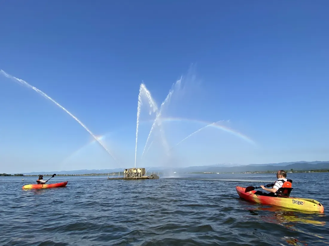 柴山潟をカヤックで進む人と、湖上の噴水にかかる虹
