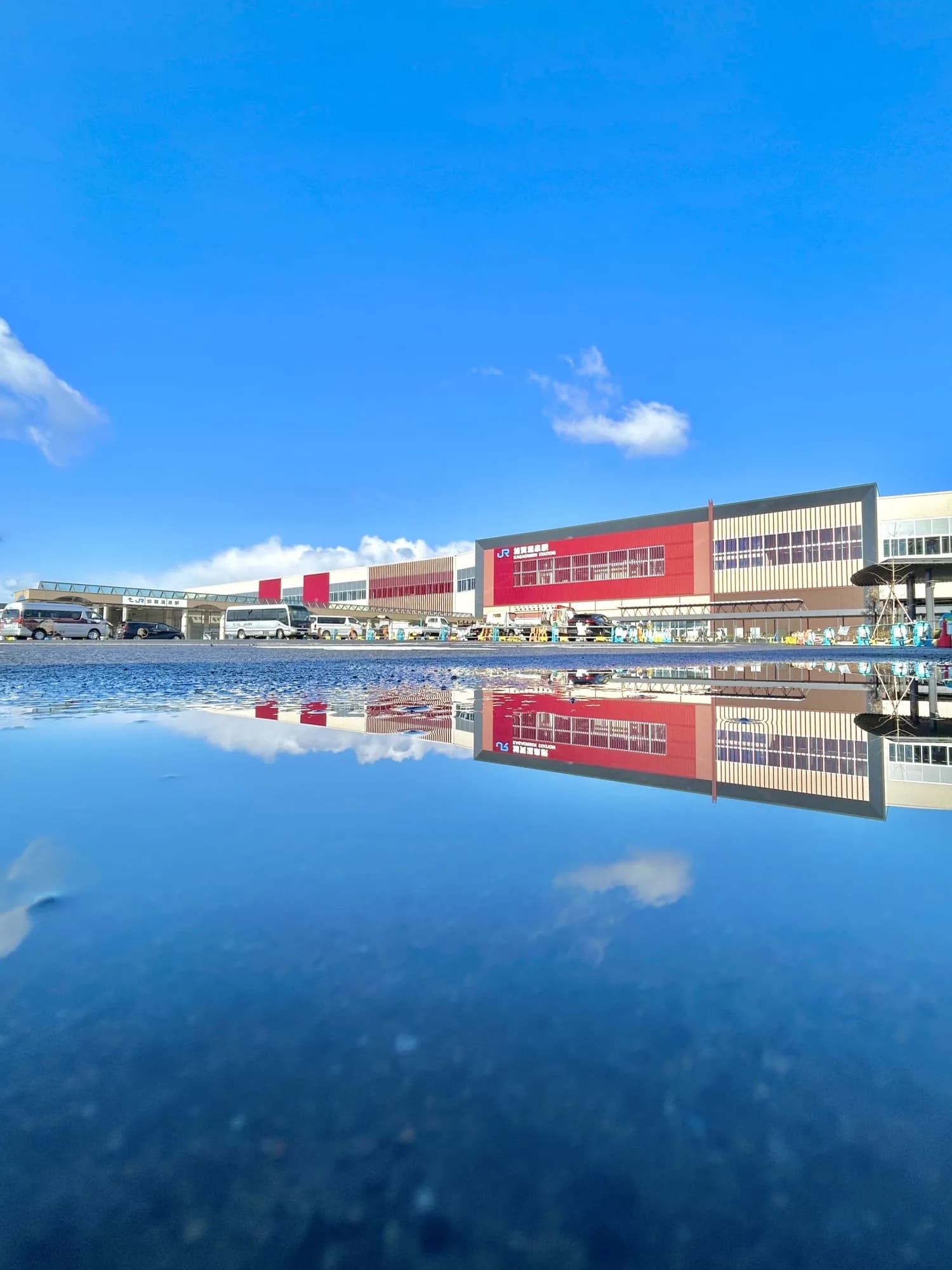 JR Kaga Onsen Station on the Hokuriku Shinkansen — red facade and blue sky reflected in a puddle